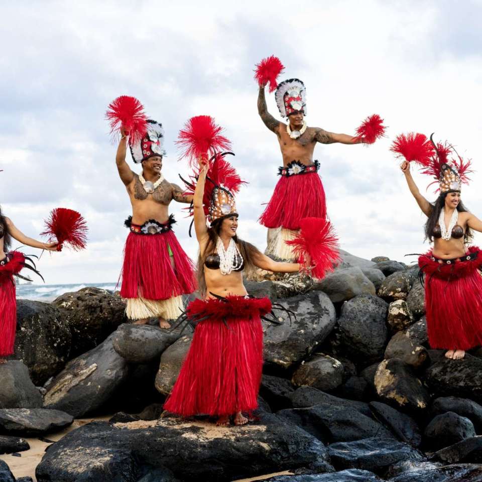Five dancers in red grass skirts and headdresses performing on rocks by the sea.