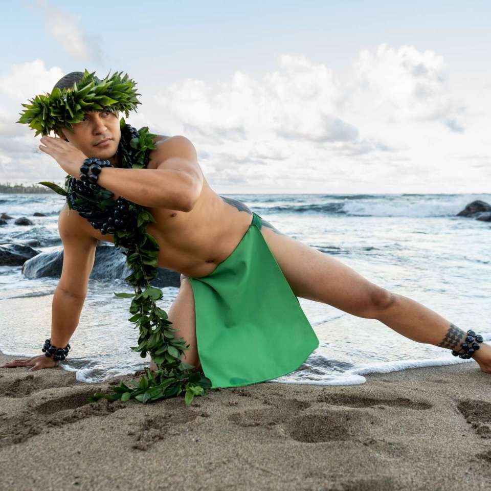 Man in green attire with leaf crown posing on sandy beach by the ocean.