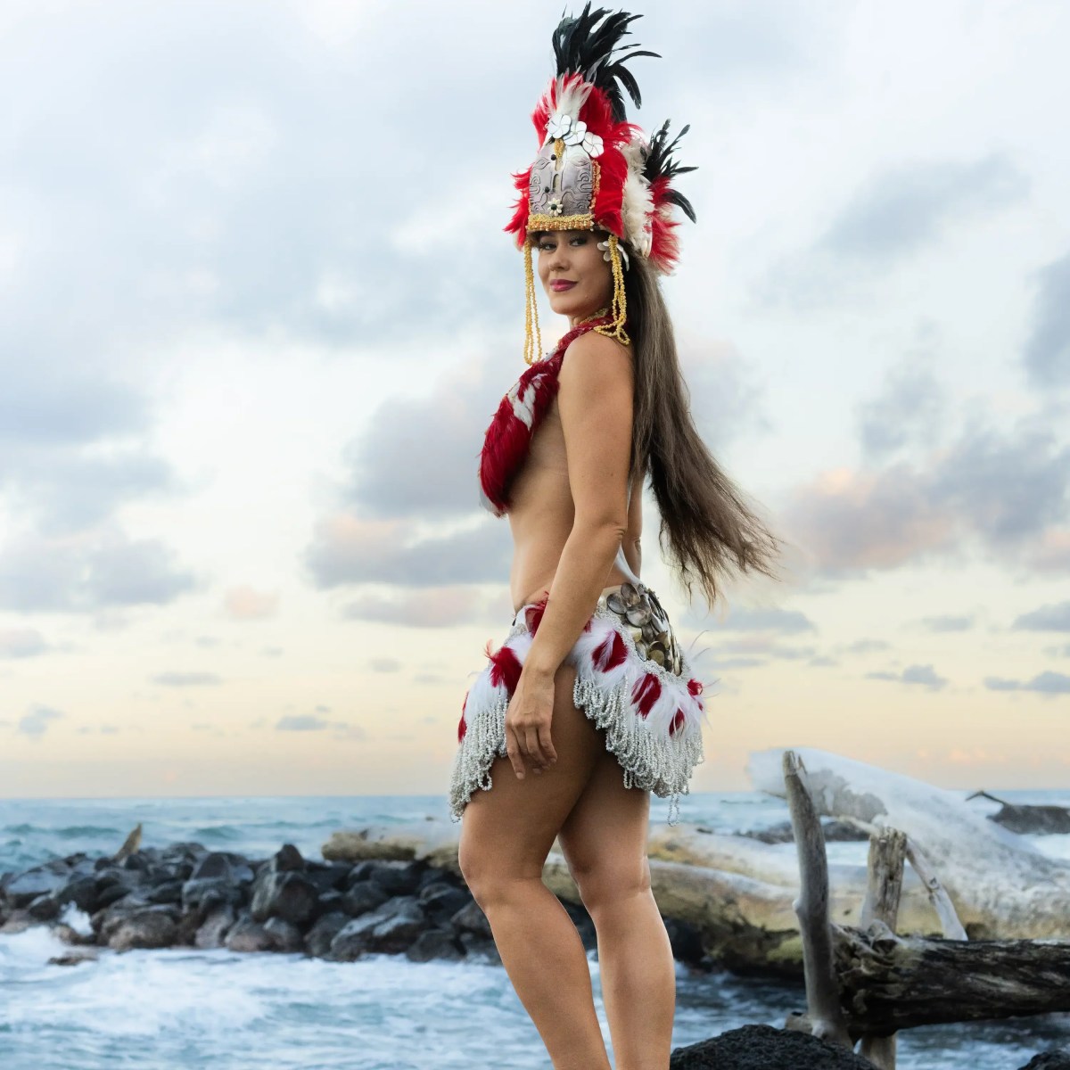 Woman in traditional costume with feathered headdress stands on rocks by the ocean.