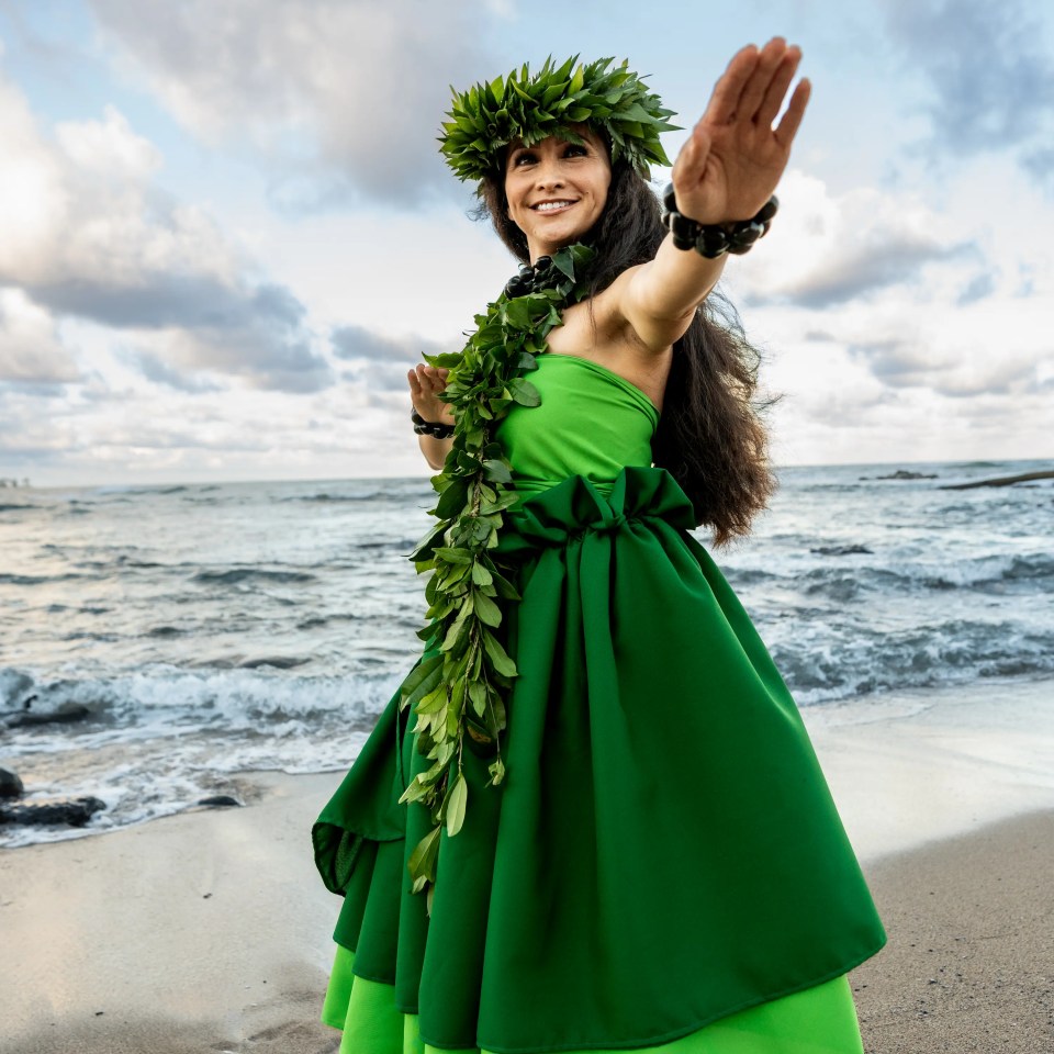 Person in green dress and leaf crown dances on beach, hand extended, with ocean and sky in background.