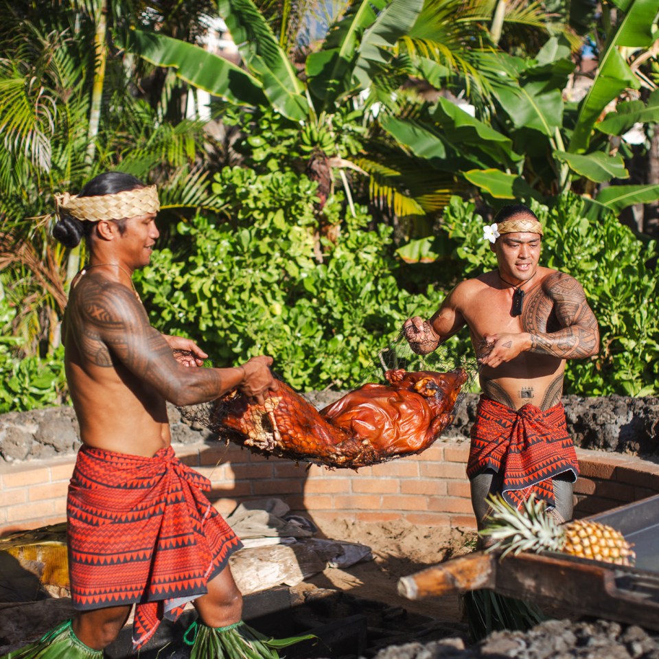 Two men in traditional attire carrying a roasted pig outdoors.